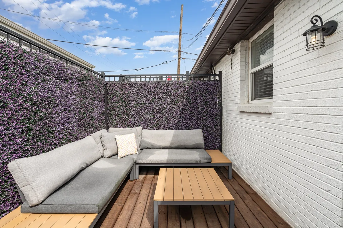 A tranquil outdoor living area with a large sectional sofa, wooden coffee table, decorative pillows, and a lush wall of greenery in the background. The space feels secluded yet open to the elements, suggesting a relaxing setting suited for conversation, reading, or unwinding after a day in the city.