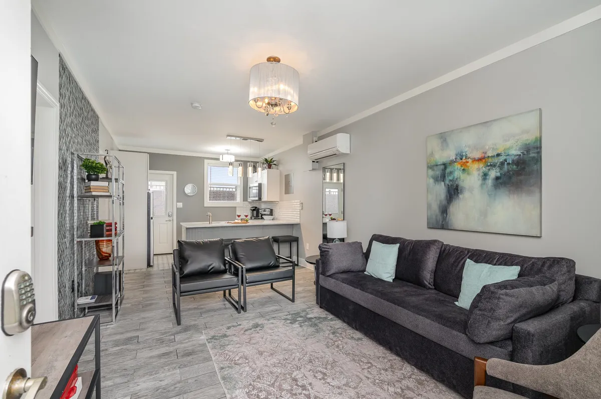 The image showcases a bright and inviting living area with a plush sofa adorned with decorative pillows, a striking abstract painting above, elegant chandelier lighting, and a seamless view into the modern open kitchen. The space is styled for both relaxation and entertaining, suggesting a comfortable and sociable atmosphere.