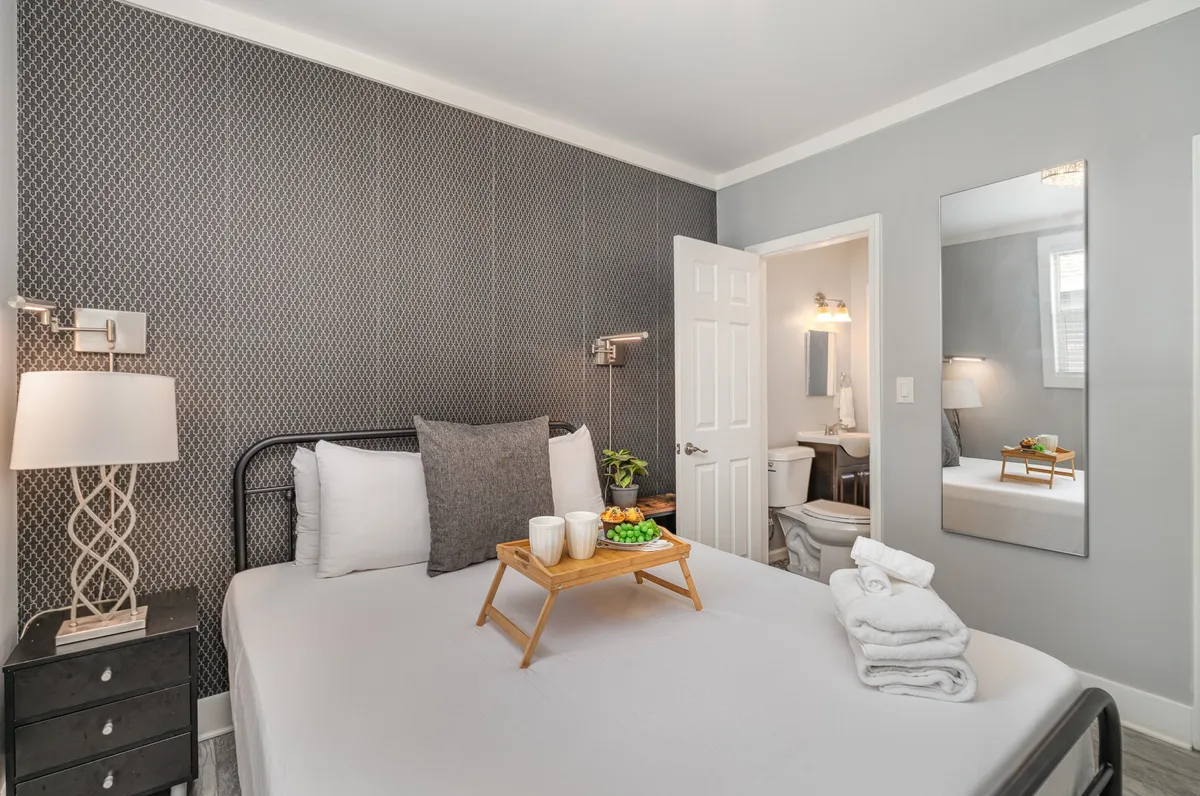 A tranquil bedroom scene featuring a neatly made queen bed with decorative pillows, a nightstand with a warm lamp, a mirror on the wall, and an open bathroom door hinting at the en suite Jack & Jill bathroom layout.