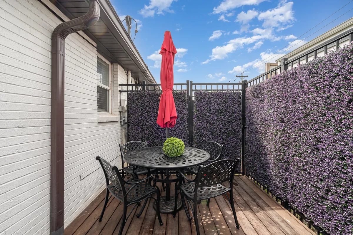 A cheerful patio scene showcasing a circular black table set for four, surrounded by black metal chairs beneath a vivid red umbrella. A green plant forms the centerpiece, and a purple floral wall adds a playful touch. The setting is sunlit and cozy, visually supporting both casual meals and celebratory occasions outdoors.
