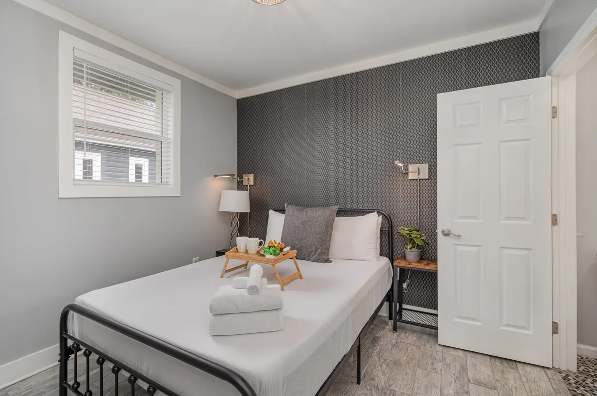 A serene bedroom with a queen bed neatly dressed in white linens, a serving tray with breakfast items atop the bed, a decorative geometric wall, and a bedside plant for a homey feel.