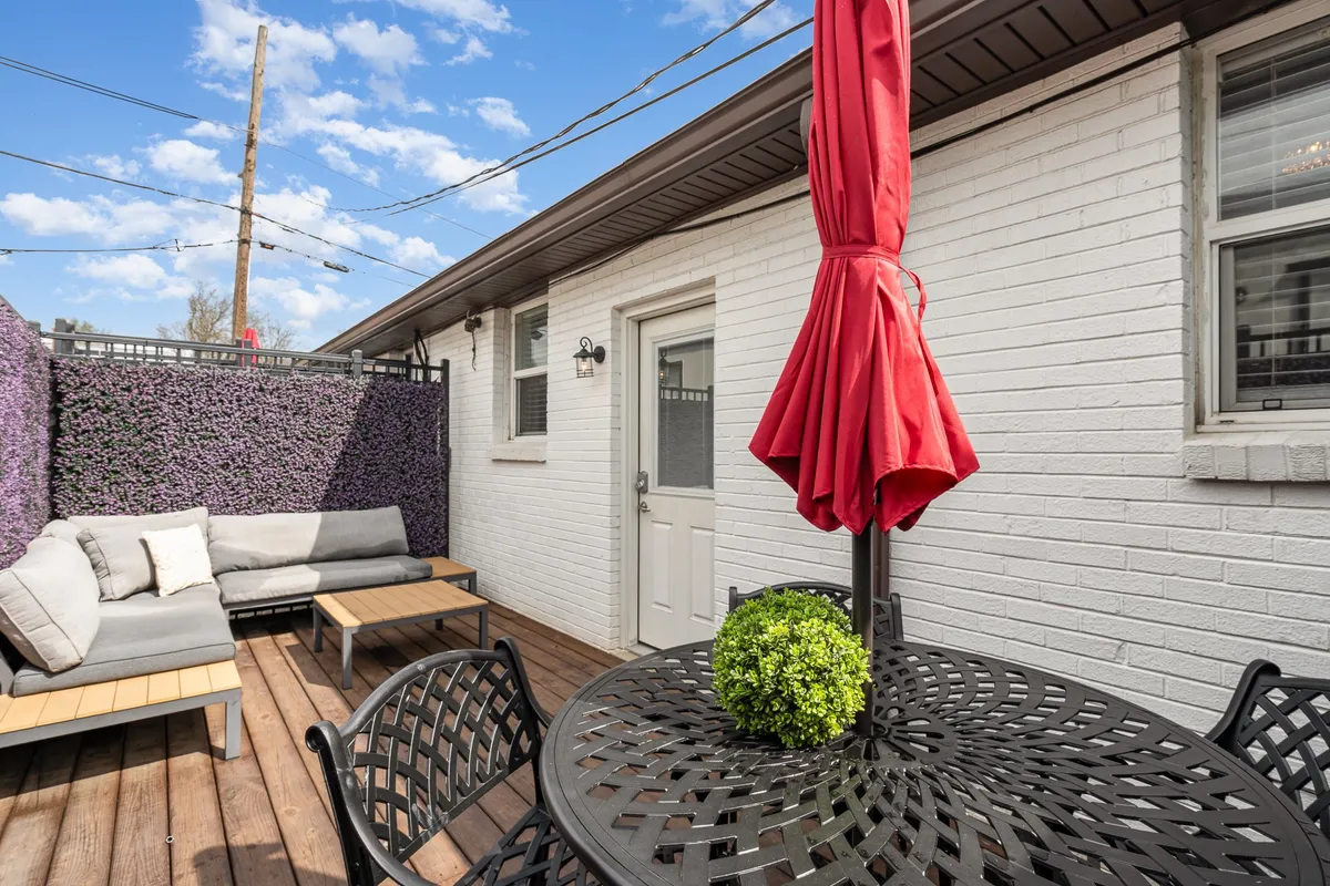 A private patio scene featuring a red umbrella providing shade over a cushioned outdoor sofa and a wooden table. Lush green plants surround the area, creating a sense of privacy and calm. The space is styled for relaxation or casual outdoor dining, with ample seating and natural elements that invite lingering in the sunshine or under the stars.